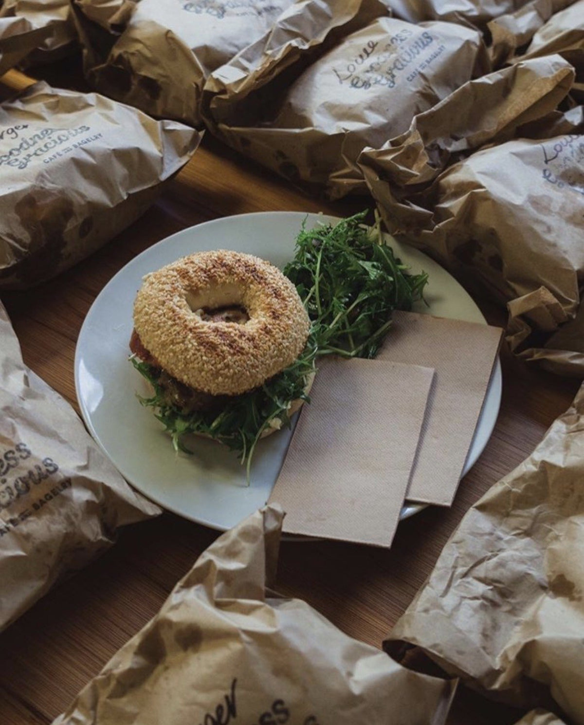 A table covered in wrapped Goodness Gracious bagels, each bag named for an order, around one plated bagel